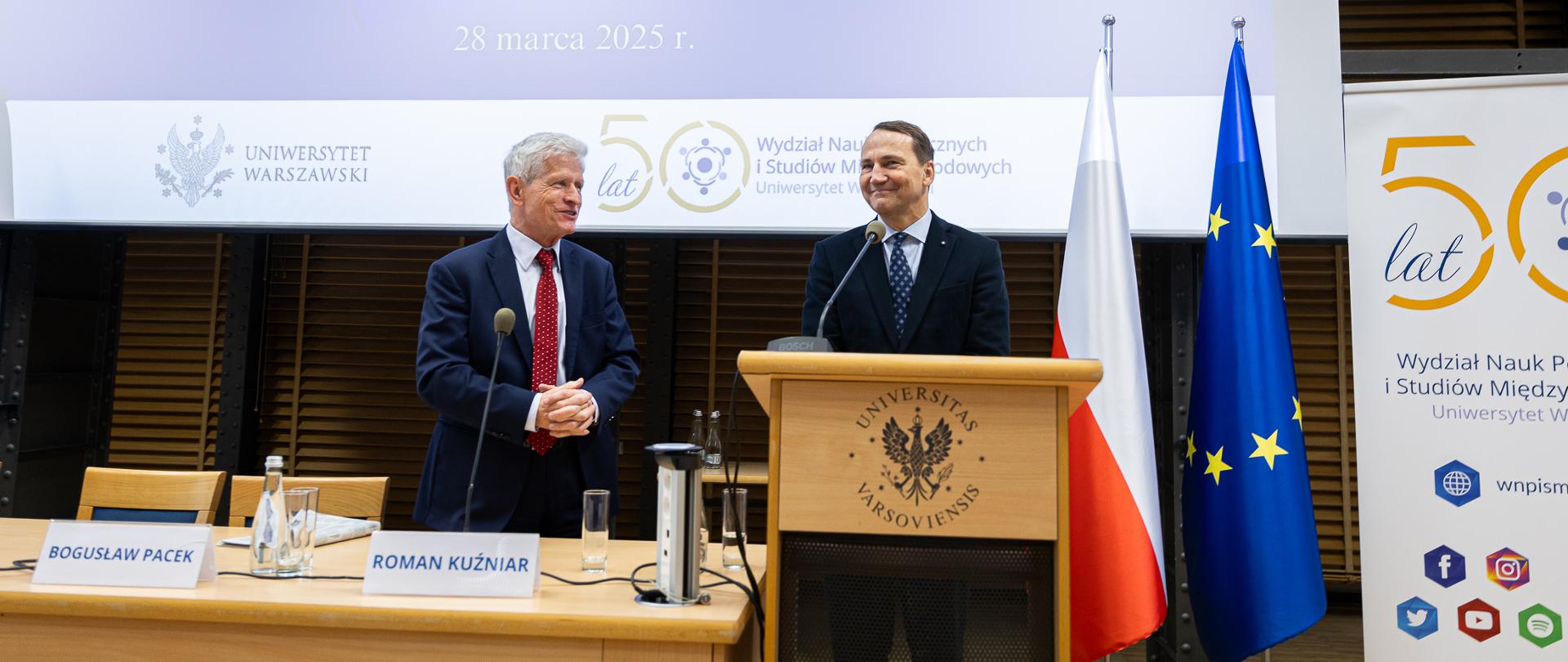 Na zdjęciu jest dwóch mężczyzn ubranych w garnitury. Za nimi są dwie flagi - Polski i Unii Europejskiej, a także ekran, na którym wyświetlana jest prezentacja. Przed nimi jest mównica oaz stół.