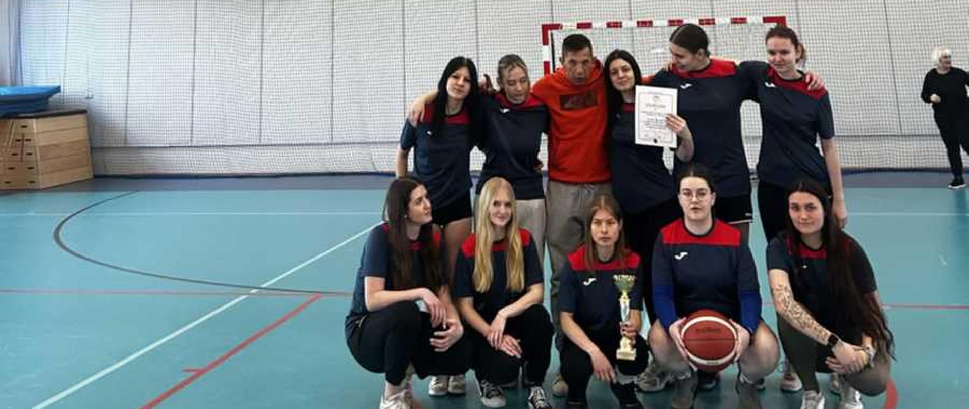 Drużyna 10-dziewcząt z trenerem Rafałem Jankowskim stoi na hali sportowej prezentując puchar i dyplom za zajęcia I miejsca w koszykówce w ramach Licealiady.