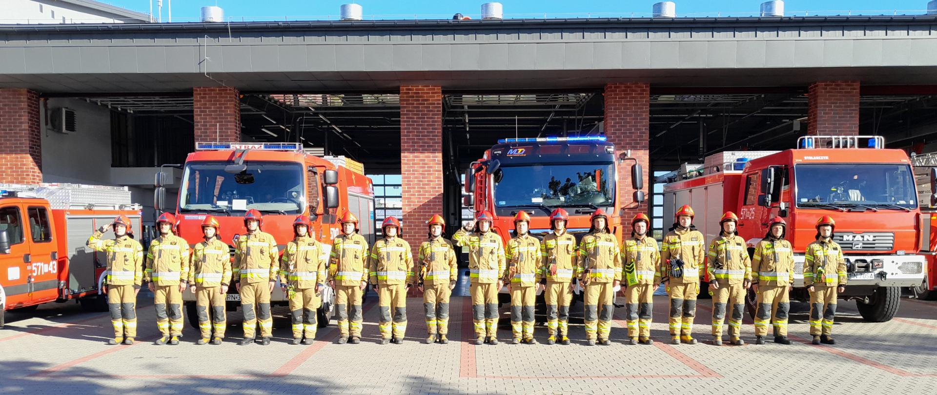 Grupa strażaków w ubraniach specjalnych koloru piaskowego, stoi w szeregu. Z tyłu, przodem ustawione są pojazdy strażackie z włączoną sygnalizacją świetlną. 