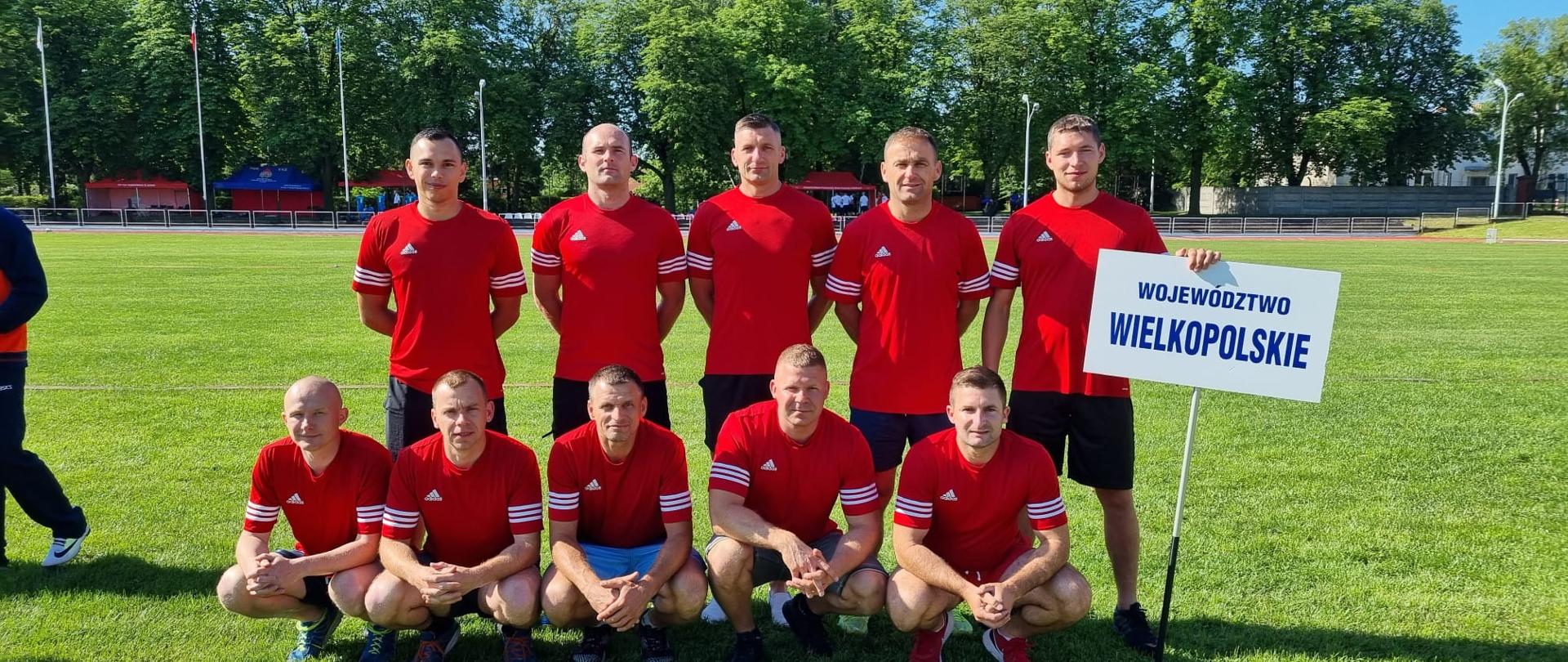 Na zdjęciu widać sportowców w czerwonych strojach sportowych. Zdjęcie zrobione na obiekcie sportowym. Za zawodnikami zielona trawa oraz tor. Zdjęcie zrobione w ciągu dnia.