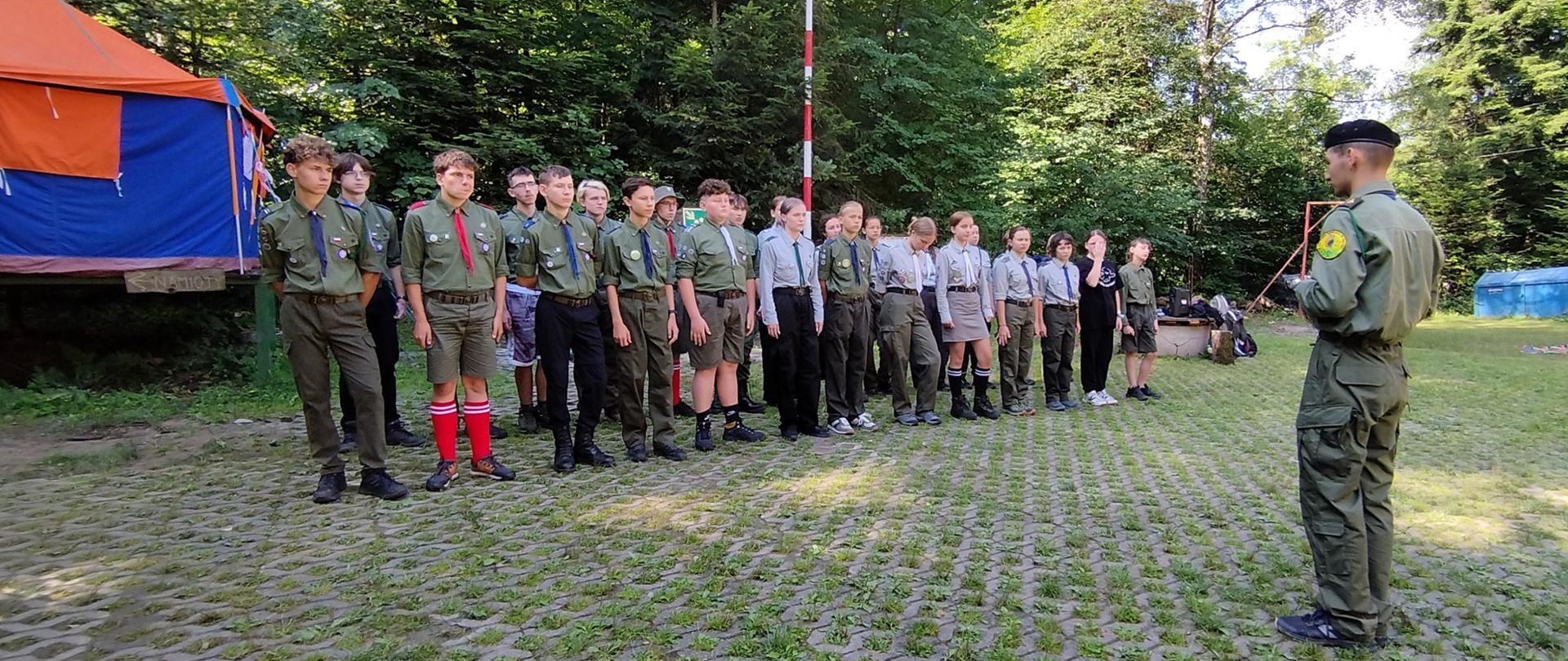 Zdjęcie grupowe. Na pierwszym planie po prawej stronie stoi harcerz (widoczny od tyłu). Na drugim planie, przodem, pododdział harcerzy w dwuszeregu. Po lewej stronie kolorowy namiot.