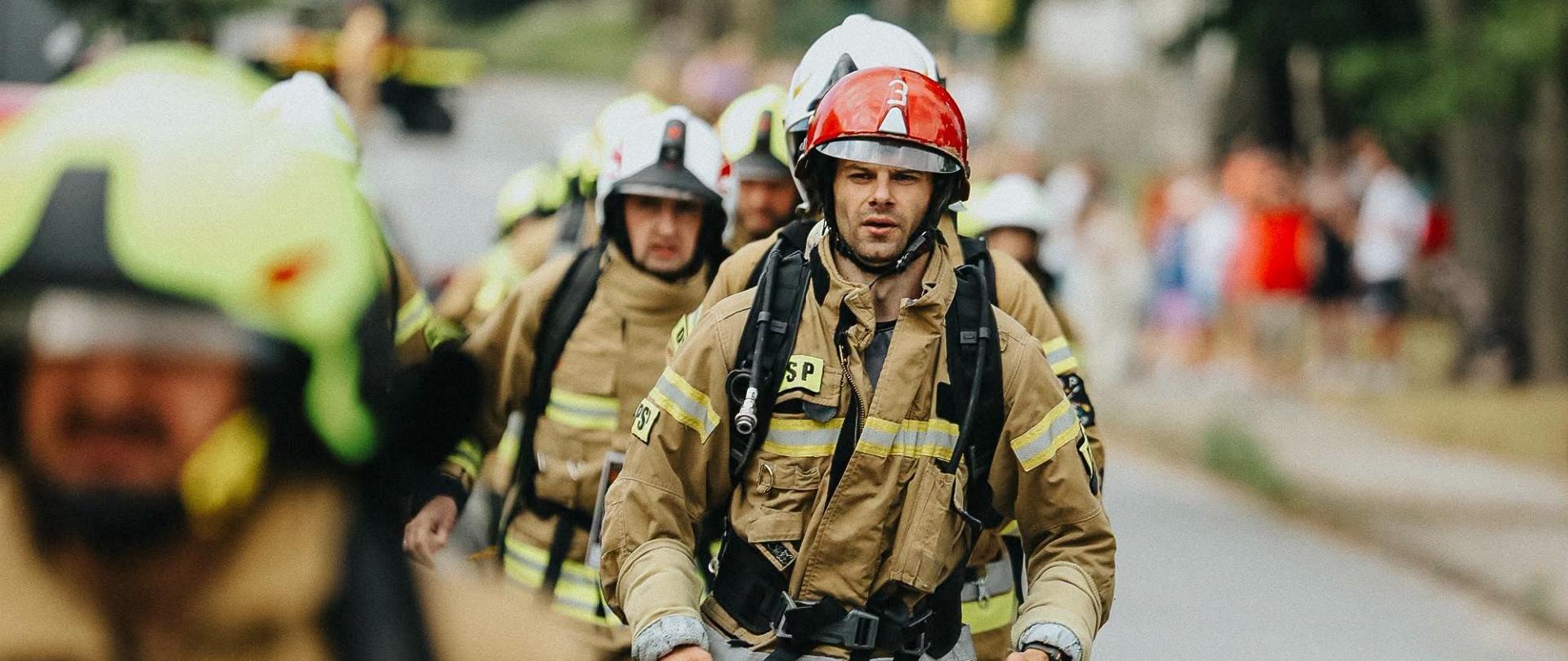 Strażacy w ubraniach specjalnych i hełmach biegną ulicą podczas wydarzenia sportowego. Na pierwszym planie widoczny jest uczestnik z numerem startowym 28, a w tle pozostali strażacy oraz kibice obserwujący bieg.