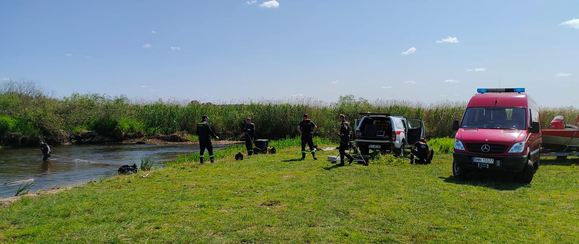 Strażacy stoją nad rzeką. Z prawej pojazdy PSP oraz łódź. Z lewej płynąca rzeka oraz zarośla.