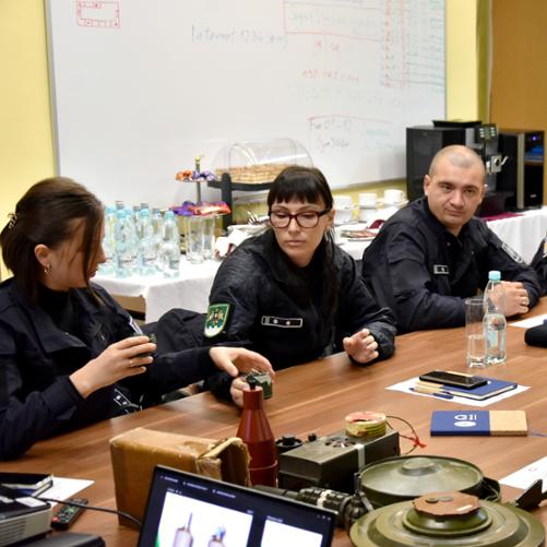 Uczestnicy szkolenia siedzą przy stole, na którym leżą materiały szkoleniowe, w tym miny i granaty