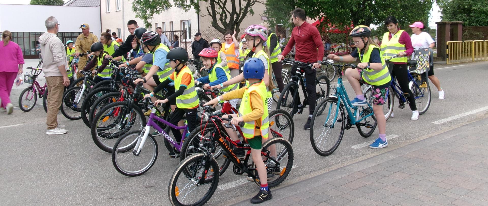 Grupa uczestników rajdu ustawiona na linii startu. Dzieci perfekcyjnie przygotowane pod względem bezpieczeństwa, w kaskach i kamizelkach odblaskowych, ustawiają rowery wraz z opiekunami, którzy będą towarzyszyć im na trasie. Na ich twarzach maluje się determinacja i pełna gotowość do startu.