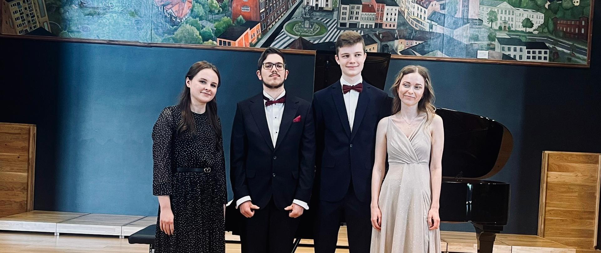 Kolorowe zdjęcie, na który stoją na scenie, w jednej linii, 4 dyplomanci na tle fortepianu i czarnej ściany z dużym kolorowym obrazem przedstawiającym najważniejsze budynki i miejsca w Rybniku. Od lewej: Agnieszka Papiorek, Franciszek Elsner, Tymoteusz Opiela, Agnieszka Malec