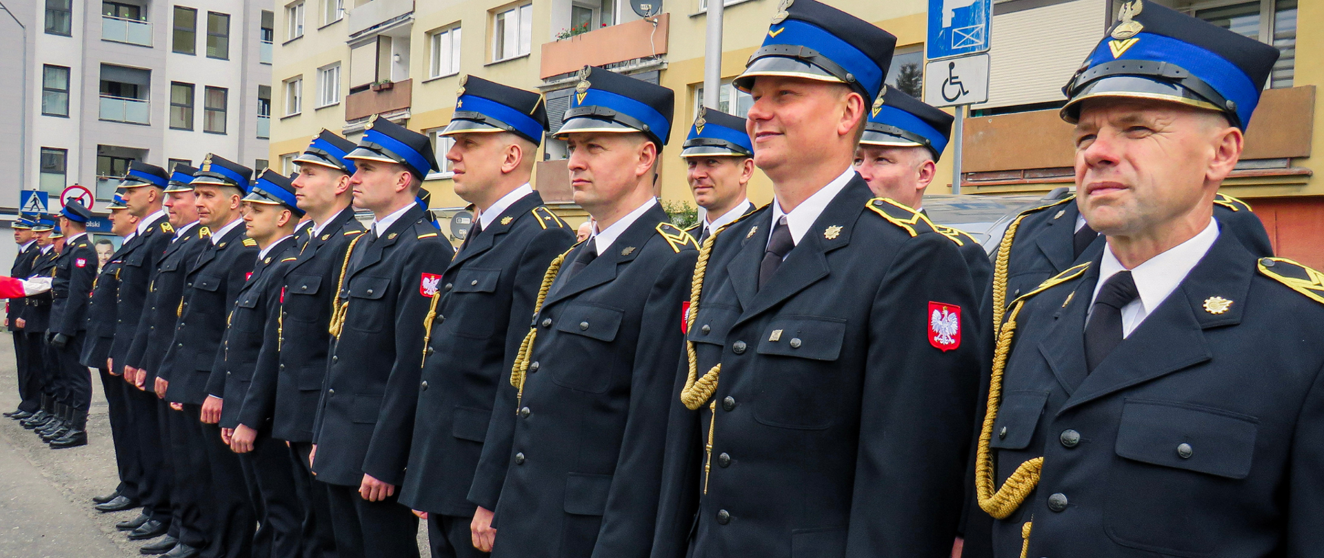 Pododdział Państwowej Straży Pożarnej stojący w dwuszeregu z pocztem flagowym na czele. 
