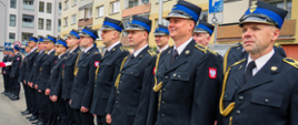 Pododdział Państwowej Straży Pożarnej stojący w dwuszeregu z pocztem flagowym na czele. 