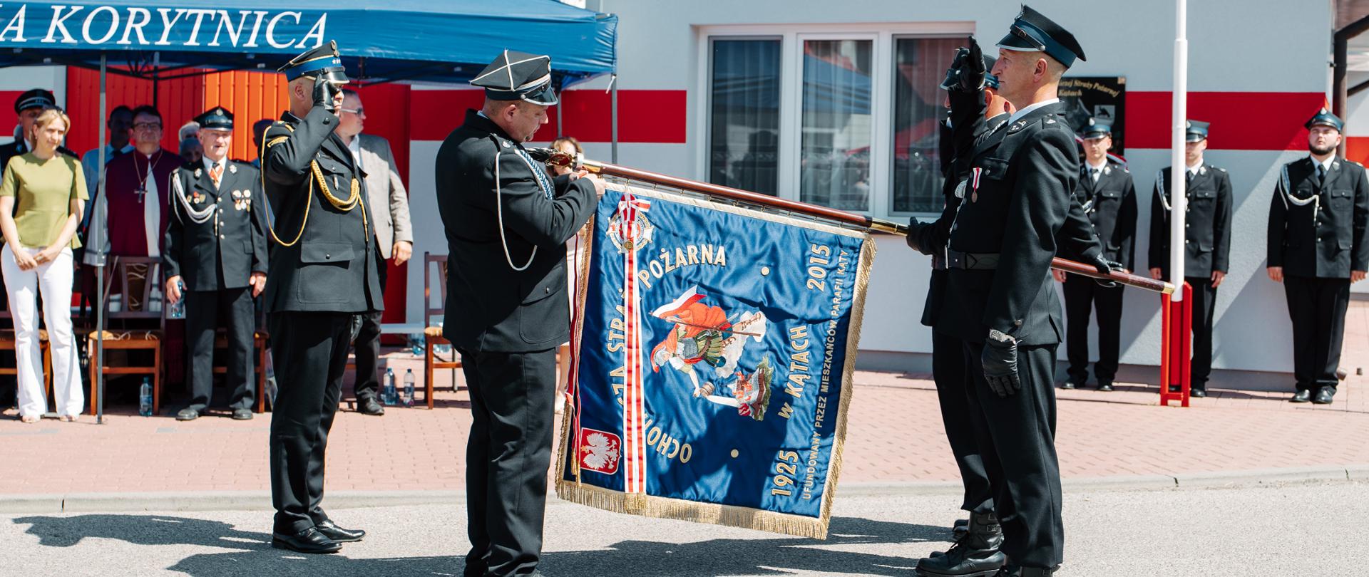 Strażacy w mundurach galowych prezentują ozdobny sztandar jednostki, a obok nich Zastępca Komendanta stoi na baczność w pozie pełnej szacunku.
