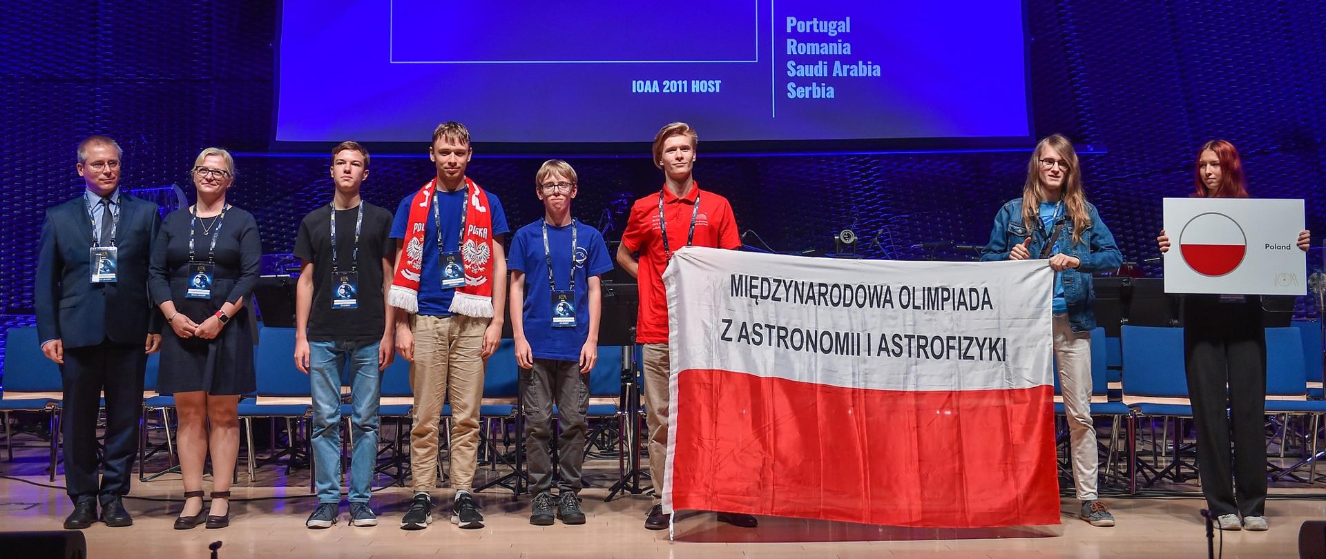 Grupa młodych ludzi stoi na scenie i trzyma flagę Polski. W tle na ekranie wyświetlana jest flaga Polski.