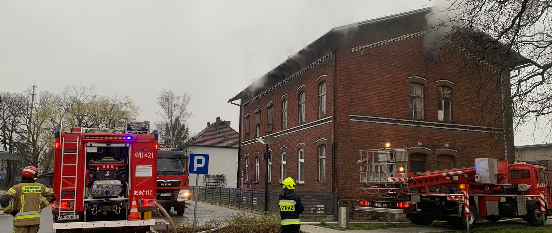 W tle budynek z czerwonej cegły z którego przez otwory okienne na poddaszu wydobywa się biały dym i para. Przed nim po prawej stronie ustawiony jest podnośnik pożarniczy ze złożonym ramieniem wysięgnika. Po lewej dwa samochody pożarnicze, jeden ustawiony tyłem drugi przodem do patrzącego. Przed samochodem ustawionym tyłem strażak PSP w piaskowego koloru ubraniu specjalnym nadzoruje prace pompy. Z auta do budynku poprowadzone są węże. W środku planu patrząc w kierunku budynku stoi tyłem strażak OSP w granatowym ubraniu specjalnym. 