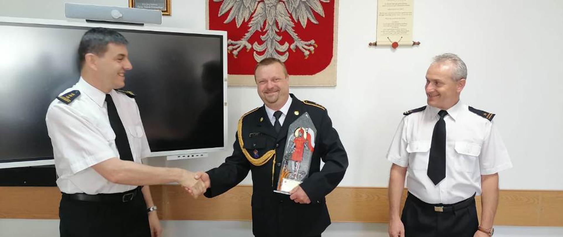Kolorowa fotografia przedstawia strażaków w świetlicy. Na pierwszym planie stoją trzej strażacy, jeden pośrodku w umundurowaniu galowym ze sznurem, a dwaj po bokach w białych koszulach z krótkim rękawem, bez nakrycia głowy. Od lewej starszy brygadier Krzysztof Folta, podaje rękę aspirantowi sztabowemu Adam Toczek. Po prawej obok stoi młodszy brygadier Bogdan Biedka. W tle jasne ściany świetlicy, pośrodku gobelin z Białym Orłem na czerwonym tle, obrazy, dyplomy i panel multimedialny.