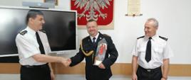 Kolorowa fotografia przedstawia strażaków w świetlicy. Na pierwszym planie stoją trzej strażacy, jeden pośrodku w umundurowaniu galowym ze sznurem, a dwaj po bokach w białych koszulach z krótkim rękawem, bez nakrycia głowy. Od lewej starszy brygadier Krzysztof Folta, podaje rękę aspirantowi sztabowemu Adam Toczek. Po prawej obok stoi młodszy brygadier Bogdan Biedka. W tle jasne ściany świetlicy, pośrodku gobelin z Białym Orłem na czerwonym tle, obrazy, dyplomy i panel multimedialny.