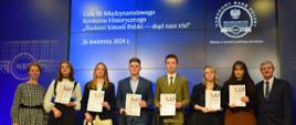 Zdjęcie grupowe z uroczystości rozdania nagród. Na scenie stoi sześcioro uczniów. W rękach trzymają dyplomy i papierowe torby z podarunkami. Wśród nich znajduje się Weronika Franiewska i Weronika Brzana. Po ich lewej stronie stoi kobieta, po prawej mężczyzna. W niebieskim tle biały napis "Gala III Międzynarodowym Konkursie Historycznym "Śladami historii Polski - skąd nasz ród", białe dwa loga Narodowego Banku Polskiego z białym napisem "Dbamy o wartość polskiego pieniądza". 