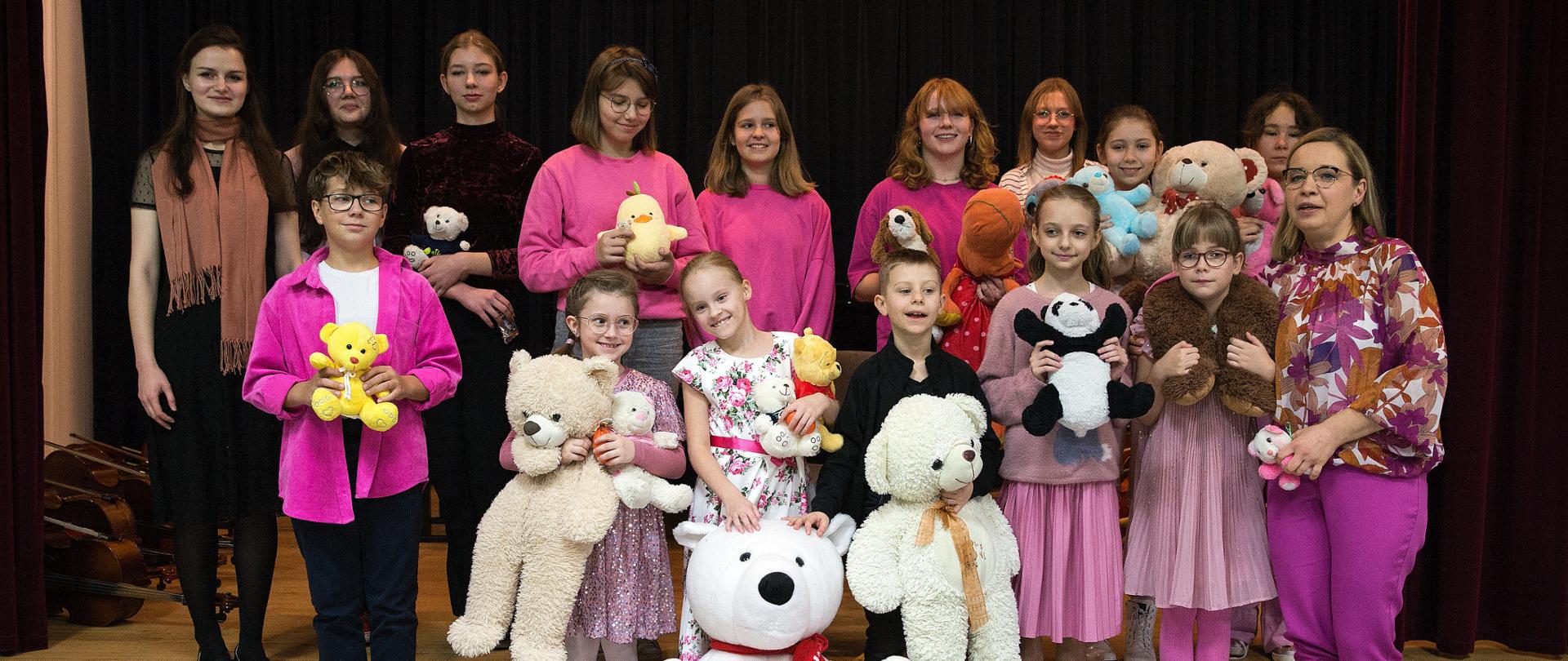Zdjęcie przedstawia ubranych na różowo uczniów, stojących na scenie z pluszowymi misiami.