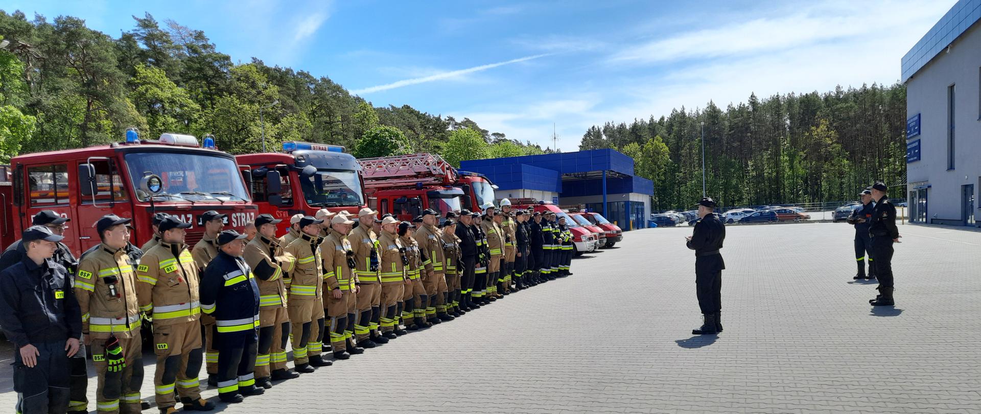 Zdjęcie wykonano w ciągu dnia przed budynkami hali fabryki mebli Gabi-bis. Po lewej stronie stoją w szeregu strażacy ubrani w ubrania specjalne, za nimi samochody ratowniczo- gaśnicze. Po prawej stoją komendant powiatowy oraz dowódca jednostki ratowniczo- gaśniczej w Sępólnie Krajeńskim. W tle budynek fabryki mebli Gabi bis. 
