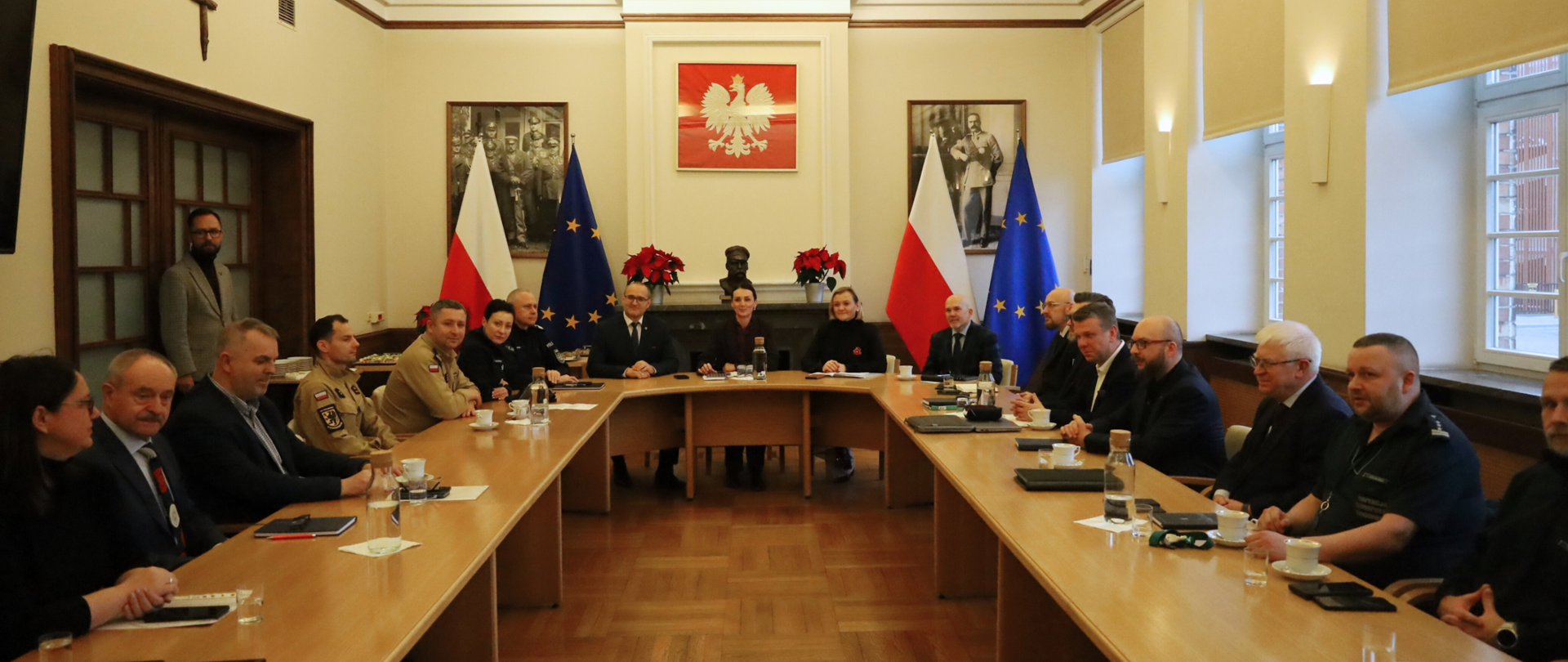 Kilkanaście osób siedzi przy okrągłym stole, na ścianie godło i zdjęcia marszałka Piłsudskiego