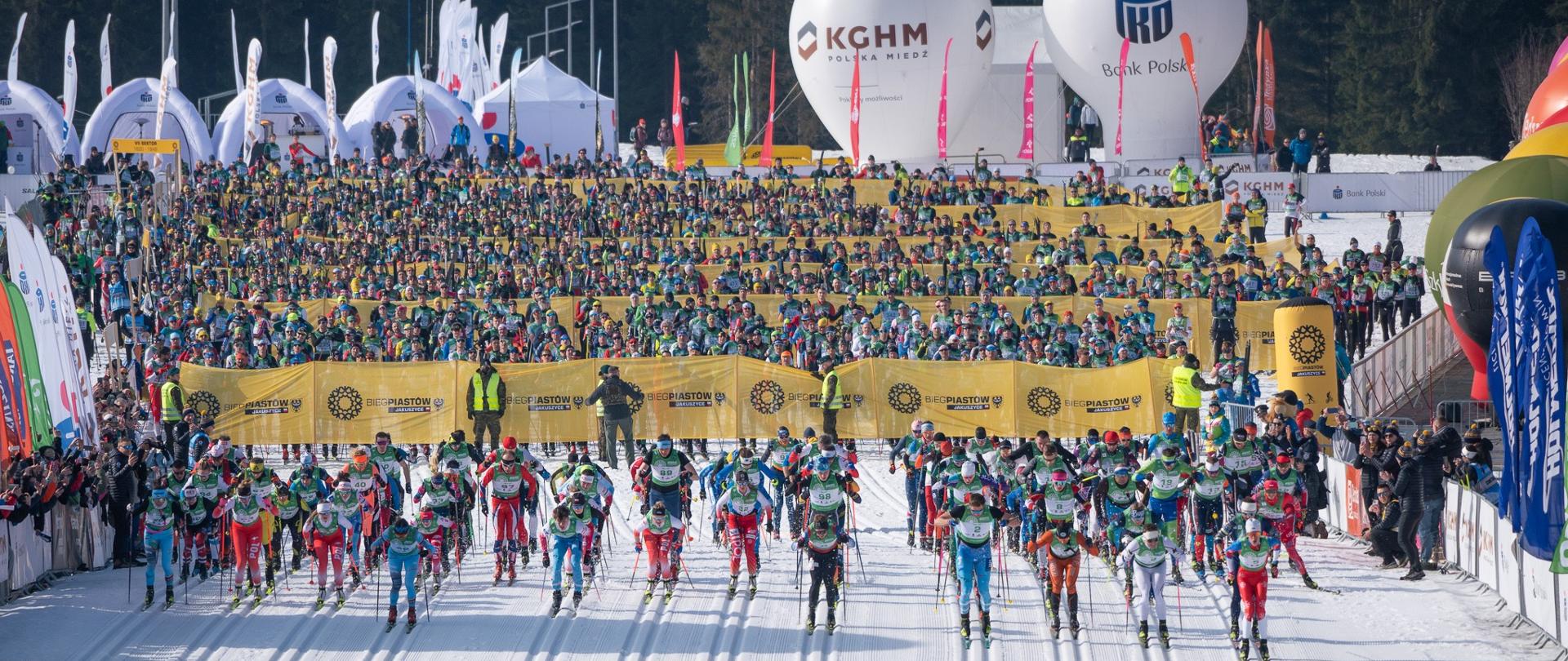 Tłum narciarzy na linii startu Biegu Piastów. Zawodnicy przygotowują się do wyścigu, stojąc w rzędach za żółtą linią. Na niebie duże białe balony z logotypami sponsorów.