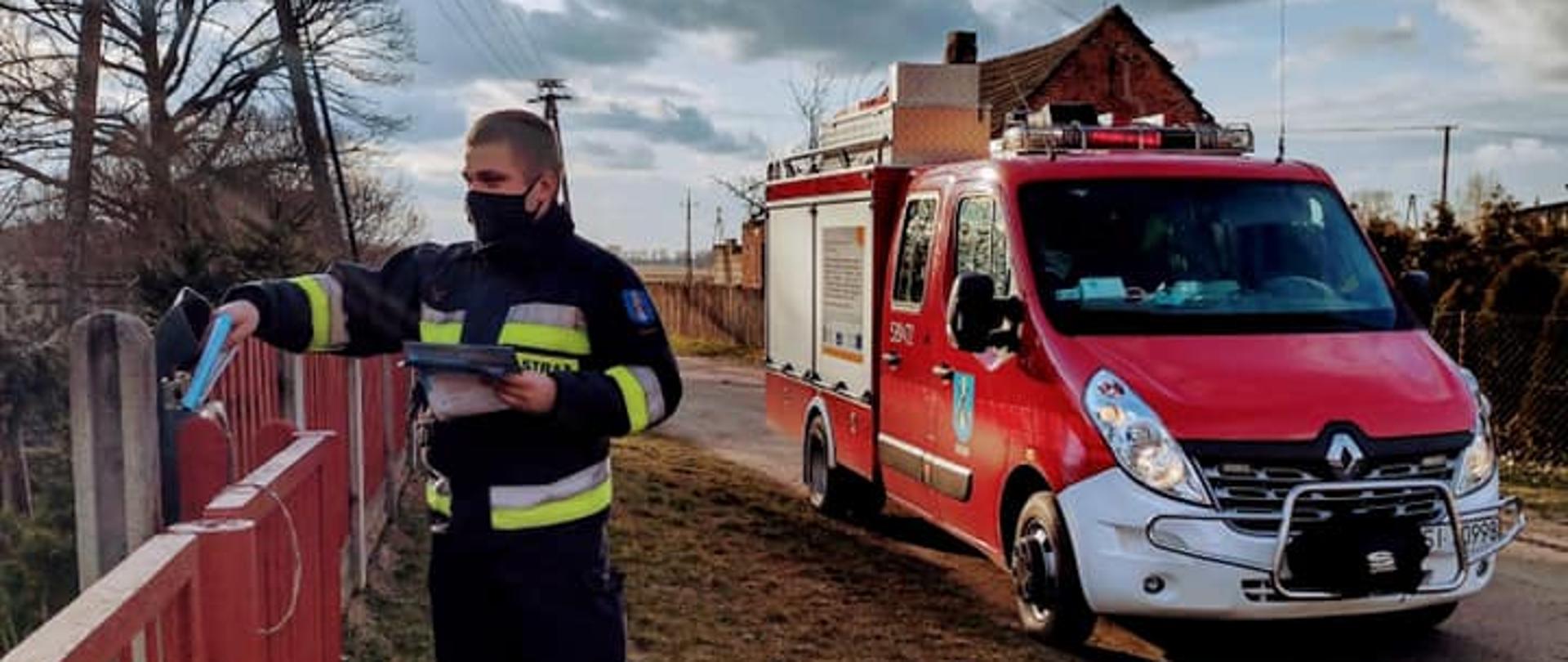 Strażak OSP wkłada do skrzynki pocztowej przy posesji ulotkę o szczepieniach. W tle samochód strazacki