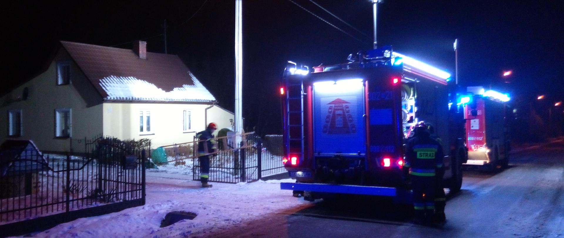 Fotografia przedstawia samochód pożarniczy na pierwszym planie, na drugim planie znajduje się budynek mieszkalny w którym doszło do pożaru. Nie widać dymu ani ognia. Strażak przy bramie wjazdowej trzyma drabinę.