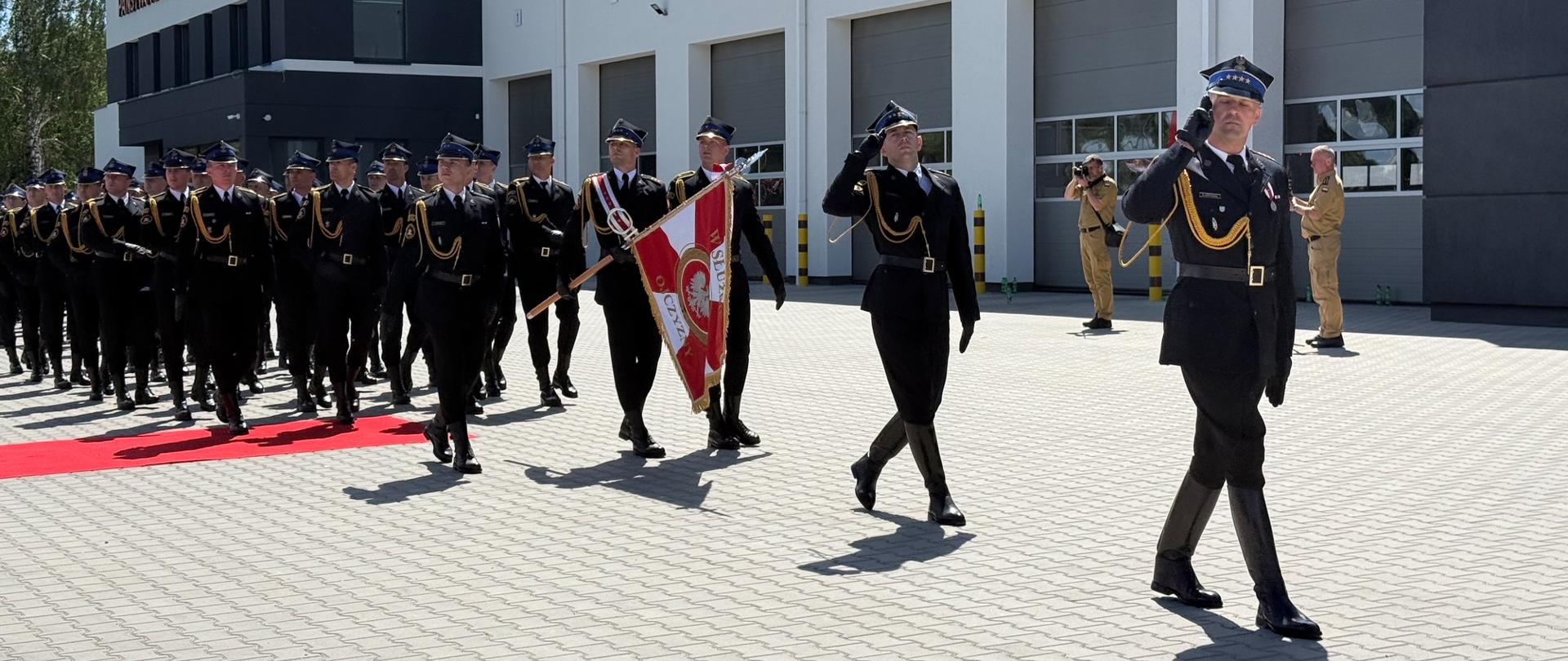 Kompania honorowa PSP maszeruje przed budynkiem jednostki. Na czele pododdziału znajduje się dowódca, za nim poczet sztandarowy niosący sztandar Państwowej Straży Pożarnej.
