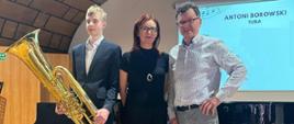 zdjęcie kolorowe. w auli koncertowej szkoły, na podwyższonej scenie stoją: uczeń z instrumentem - tubą, akompaniatorka i nauczyciel. W tle widać otwarty fortepian, z tyłu na ścianie tablica informacyjna z napisem "Antoni Borowski, tuba" (reszta napisu nieczytelna).