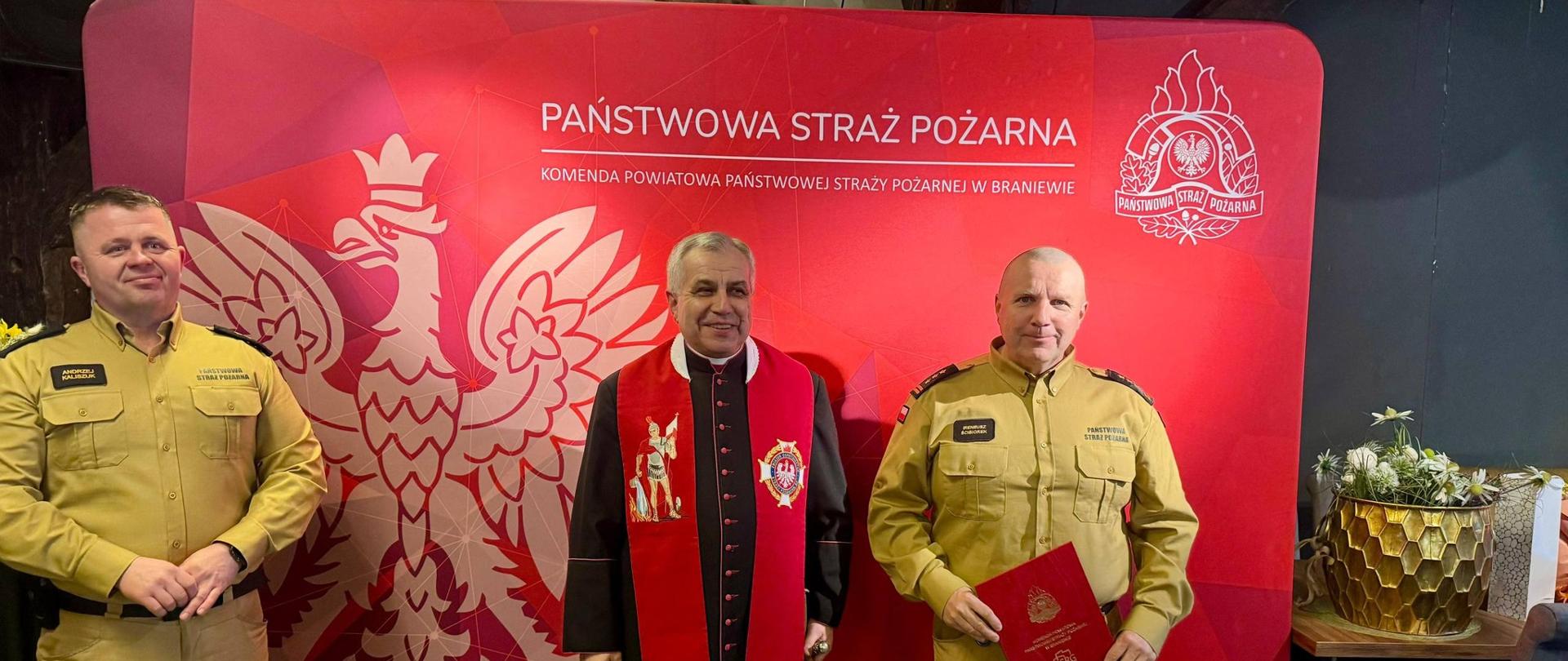 Z prawej i lewej strony strażacy w jasnych mundurach, w środku stoi ksiądz, kapelan strażaków. W tle ścianka czerwona z napisem państwowa straż pożarna i godło Polski.
