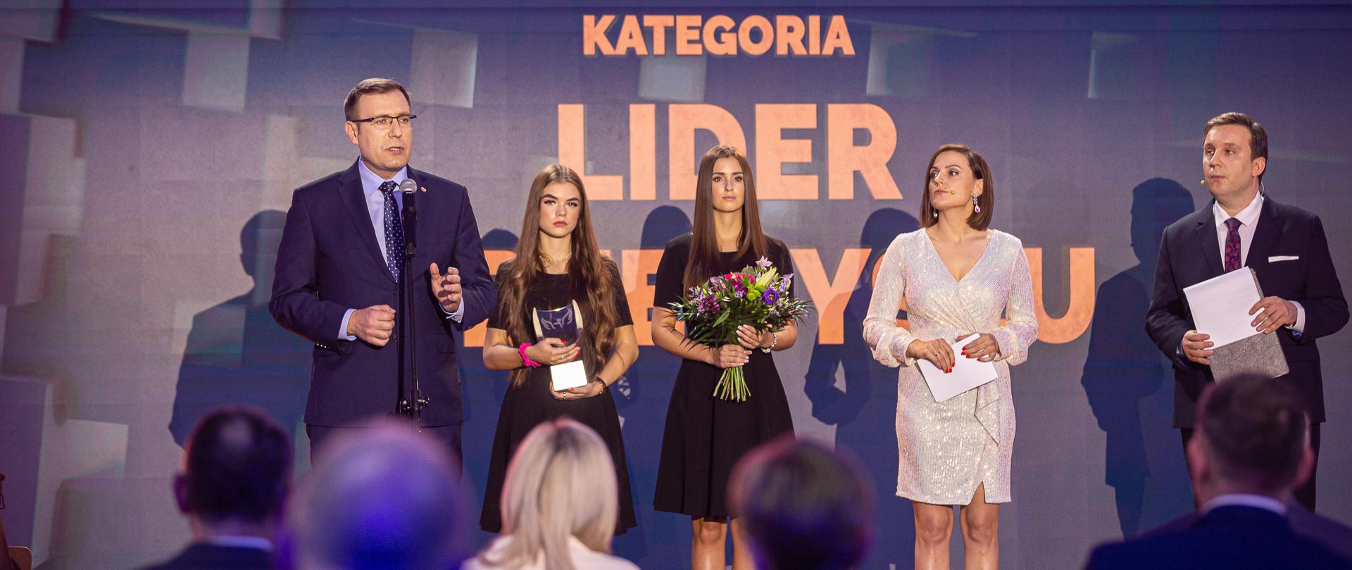 Wiceminister Maciej Małecki i prowadzący Galę, bliski plan. W tle ścianka z napisem Kategoria Lider Przemysłu. 