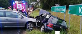 Centralny punkt zdjęcia stanowią dwa rozbite auta osobowe. W tle pojazdy Straży Pożarnej. Jest pochmurno. Jedno z aut uderzyło w bok drugiego.