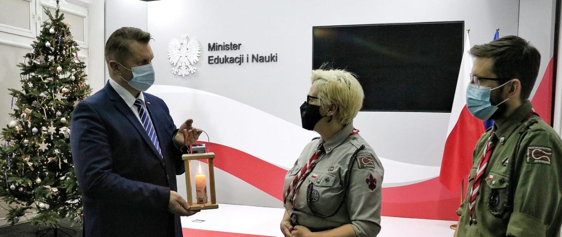 Minister Edukacji i Nauki Przemysław Czarnek otrzymujący Betlejemskie Światło Pokoju od harcerzy. W tle stoi udekorowana choinka.