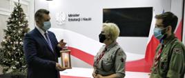 Minister Edukacji i Nauki Przemysław Czarnek otrzymujący Betlejemskie Światło Pokoju od harcerzy. W tle stoi udekorowana choinka.