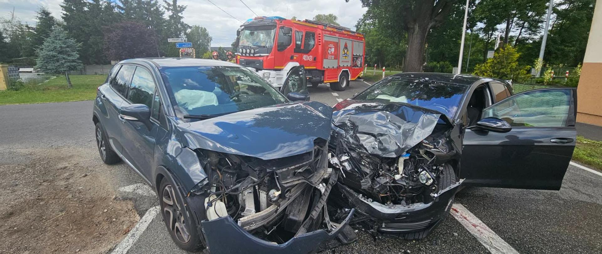 Zdjęcie przedstawia strażaków podczas wypadku drogowego.
W tle pojazdy, droga i tereny zielone.