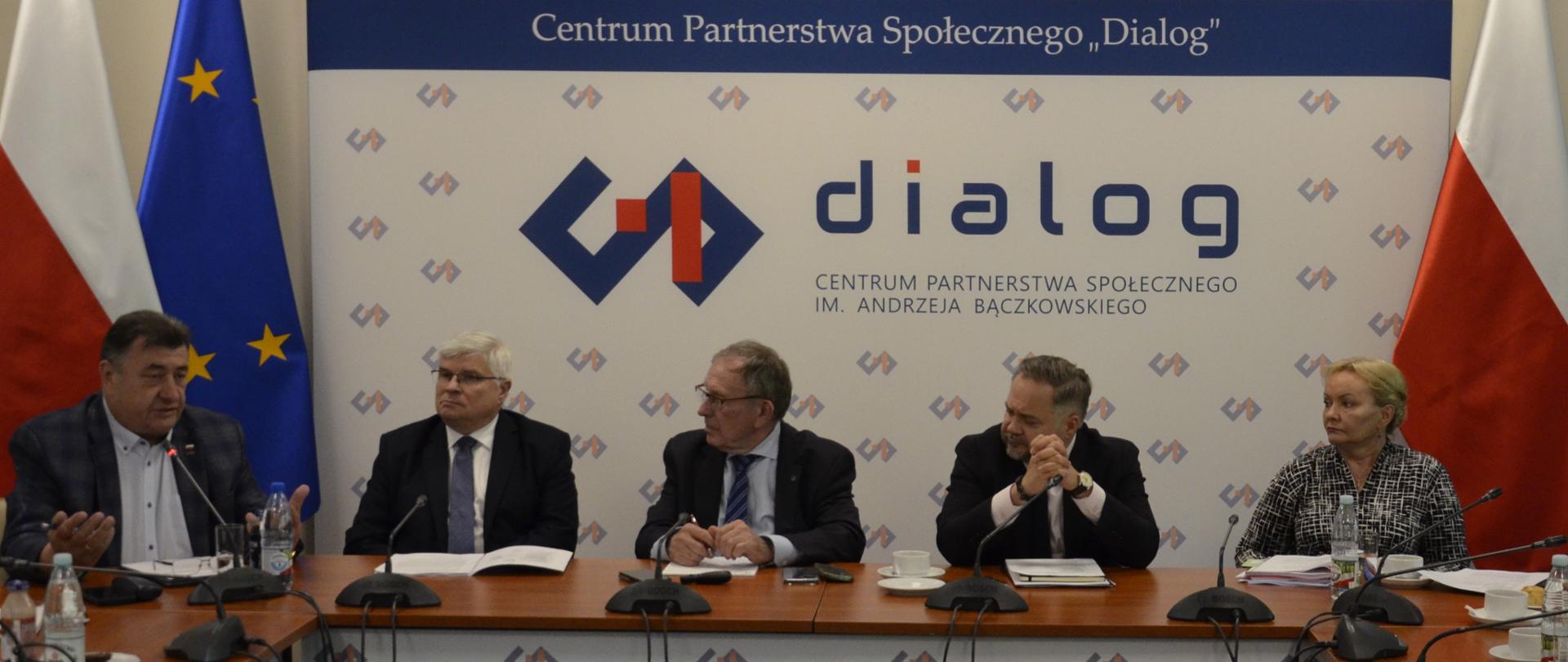 Na zdjęciu widnieje stół prezydialny za którym siedzą: Pan Maciej Lasek, Sekretarz Stanu w Ministerstwie Infrastruktury; Pan Konrad Gołota, Podsekretarz Stanu w Ministerstwie Aktywów Państwowych; Pani Małgorzata Baranowska, Podsekretarz Stanu w Ministerstwie Rodziny, Pracy i Polityki Społecznej; Pan Marek Dras, Współprzewodniczący Zespołu ze strony pracodawców oraz Pan Roman Jakim, Współprzewodniczący Zespołu ze strony związków zawodowych. W tle znajduje się baner z napisem Centrum Partnerstwa Społecznego „Dialog” im. Andrzeja Bączkowskiego. Po lewej i prawej stronie banneru znajdują się flagi Polski. Po lewej stronie znajduje się także flaga UE.
