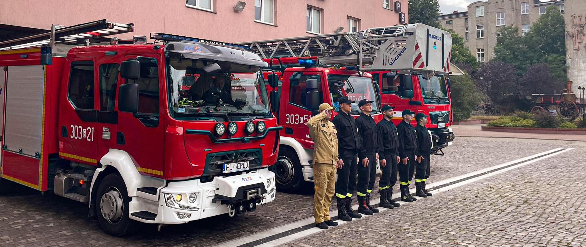 sześciu strażaków w ubraniach koszarowych stojących na zbiórce przed garażem jednostki ratowniczo-gaśniczej za strażakami garażem stoją trzy samochody gaśnicze 