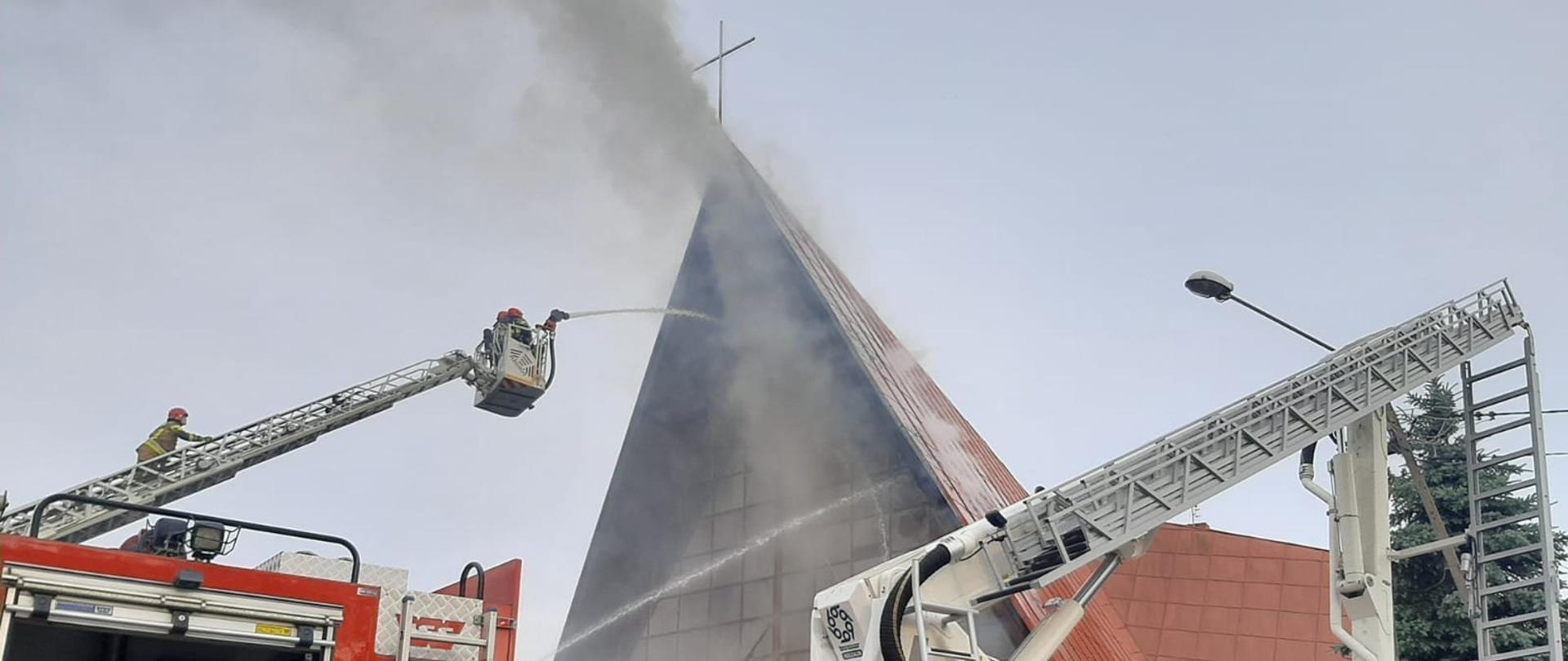 Na pierwszym planie dwa samochody pożarnicze - gaśniczy i drabina. W tle widać jak z kościoła wydobywają się kłęby szarego dymu, a strażacy podają prądy gaśnicze.
