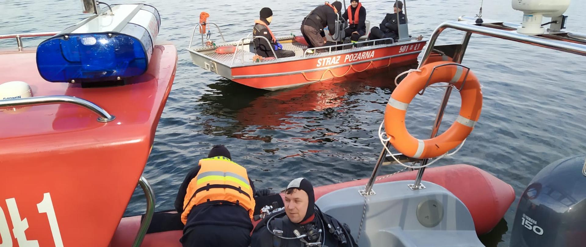 Dwie łodzie pływają po jeziorze. Na jednej sternik rozmawia z nurkiem w wodzie, drugi nurek siedzi w łodzi. Na drugiej łódce jest 4 strażaków, w tym jeden ubrany w suchy skafander do nurkowania.