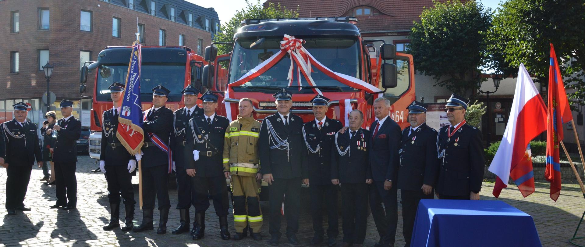 Na zdjęciu strażacy ochotnicy przed nowo otrzymanym samochodem rat. gaś. Z prawej strony stół, flagi. Z tyłu budynki mieszkalne. 