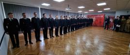Na zdęciu wewnątrz pomieszczenia ustawieni w szeregu strażacy w umundurowaniu wyjściowym przed nimi stoi Komendant Miejski Państwowej Straży Pożarnej w Częstochowie