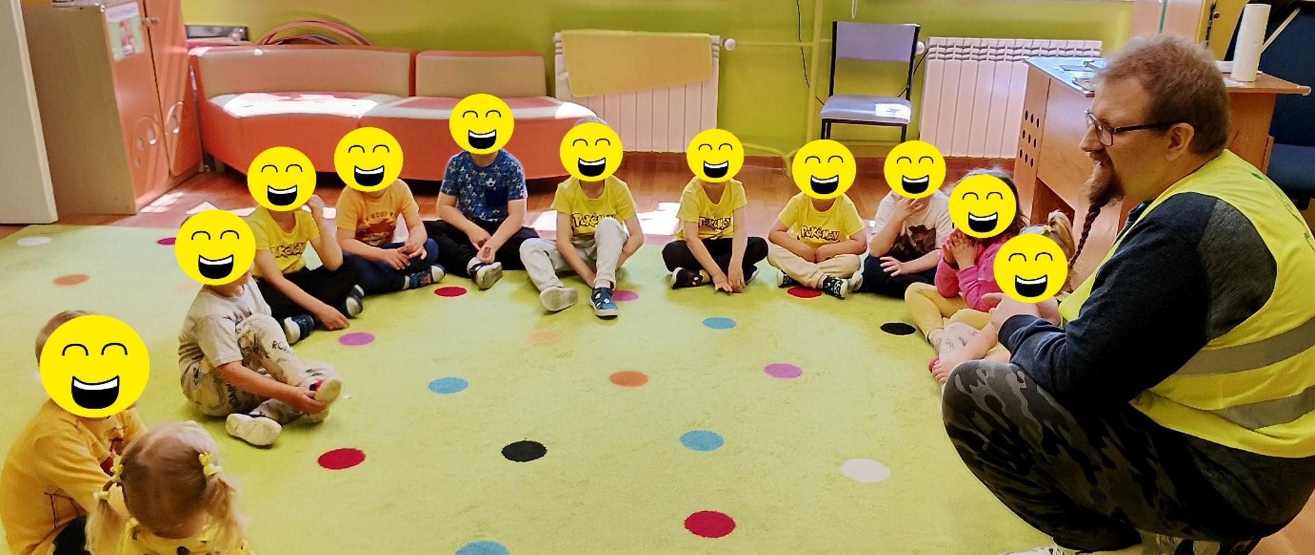 Dzieci siedzą w kręgu na jasnozielonym dywanie w przedszkolu, naprzeciwko nich kuca prowadzący zajęcia pracownik Wojewódzkiego Inspektoratu Ochrony Roślin i Nasiennictwa w Warszawie, Oddziału w Lipsku w żółtej kamizelce odblaskowej. Większość dzieci ma żółte ubrania, a sala jest kolorowo udekorowana rysunkami na oknach.