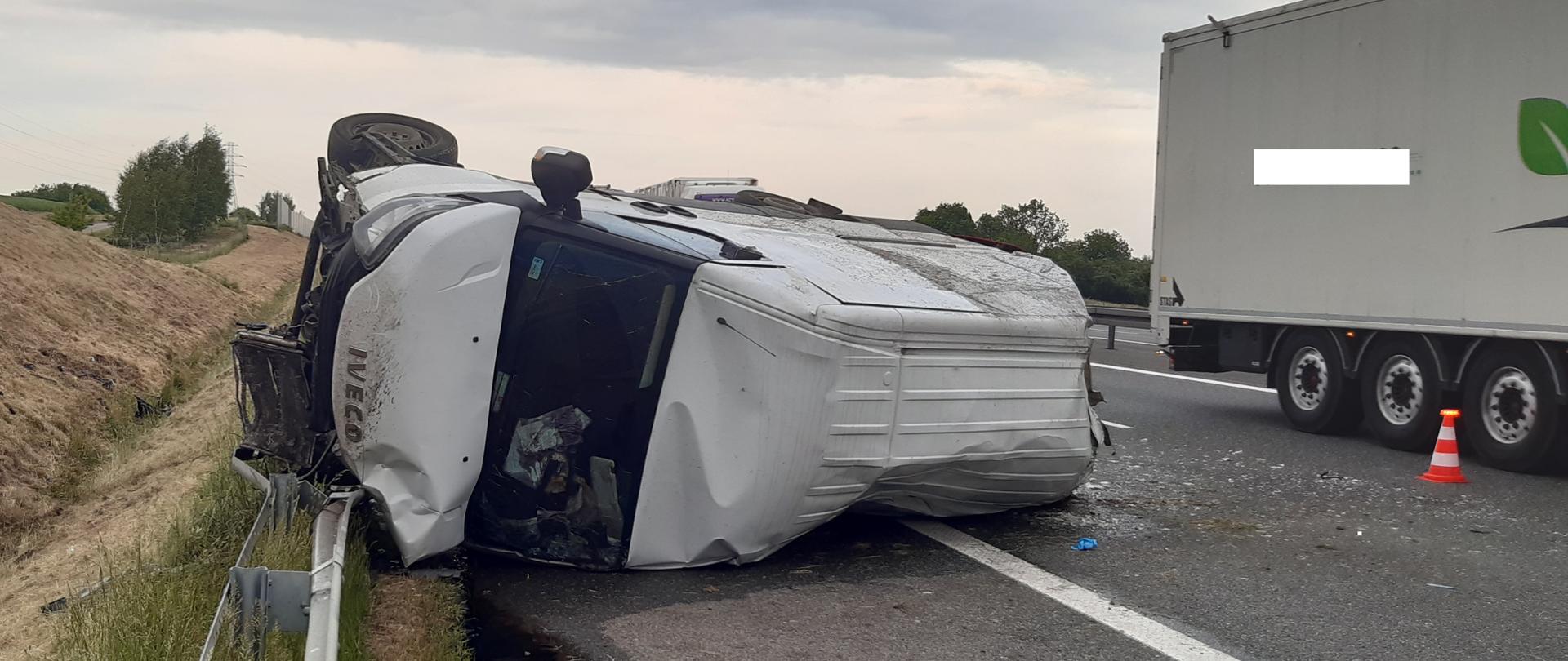 Na zdjęciu samochód dostawczy na boku po uderzeniu w bariery energochłonne. Z prawej strony naczepa od samochodu ciężarowego.