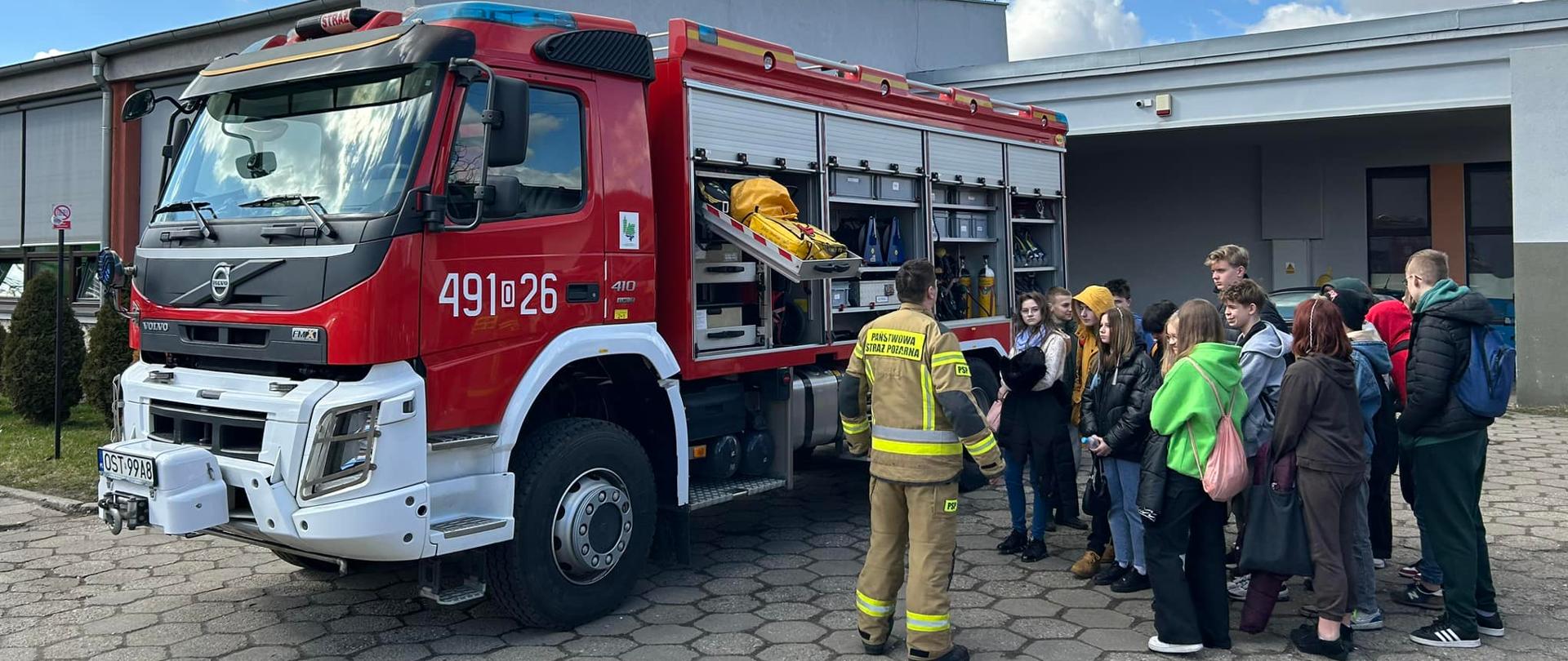 Na zdjęciu znajduje się pojazd pożarniczy z otwartymi skrytkami, strażak w ubraniu specjalnym oraz młodzież