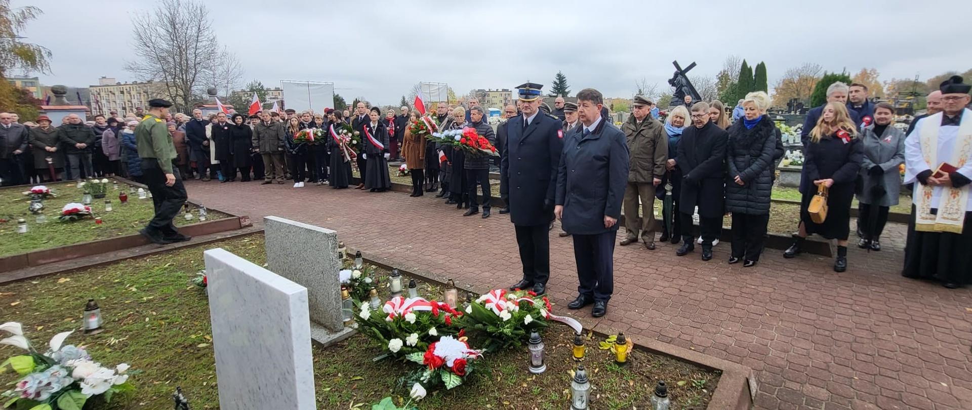 Zdjęcie przedstawia uroczystość na cmentarzu w pochmurny, jesienny dzień. Na pierwszym planie, po lewej stronie, widoczne są dwa jasne nagrobki z ułożonymi przed nimi biało-czerwonymi wieńcami, udekorowanymi wstążkami w barwach narodowych Polski. Wokół nagrobków znajdują się liczne znicze i kwiaty. W centrum kadru stoją dwaj mężczyźni w ciemnych strojach: jeden w galowym mundurze, prawdopodobnie przedstawiciel straży pożarnej, drugi w ciemnym płaszczu. Obaj stoją z powagą, patrząc przed siebie. Za nimi rozciąga się grupa ludzi, zgromadzonych wzdłuż alejki cmentarnej i na trawnikach. Wśród nich widać osoby w różnym wieku, niektóre ubrane w ciemne, formalne stroje, inne w bardziej codzienne kurtki. Niektórzy trzymają wieńce lub bukiety kwiatów. Po prawej stronie widać duchownego w szatach liturgicznych. W tle widoczne są inne nagrobki, drzewa bez liści oraz krzyż. Cała scena sugeruje obchody uroczystości patriotycznej lub rocznicowej, być może związanej z Dniem Niepodległości, mającej na celu oddanie hołdu zmarłym.