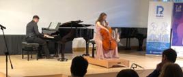 Na pierwszym planie widoczny fragment publiczności. Na scenie młoda dziewczyna gra na wiolonczeli. Za nią, przy fortepianie zasiada akompaniator.