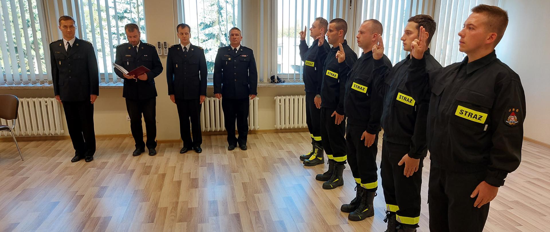 Na zdjęciu widzimy 5 strażaków składających ślubowanie, stojących w szeregu ubranych w umundurowanie koszarowe z ich prawej strony stoi 4 funkcjonariuszy tyłem do okna. 
