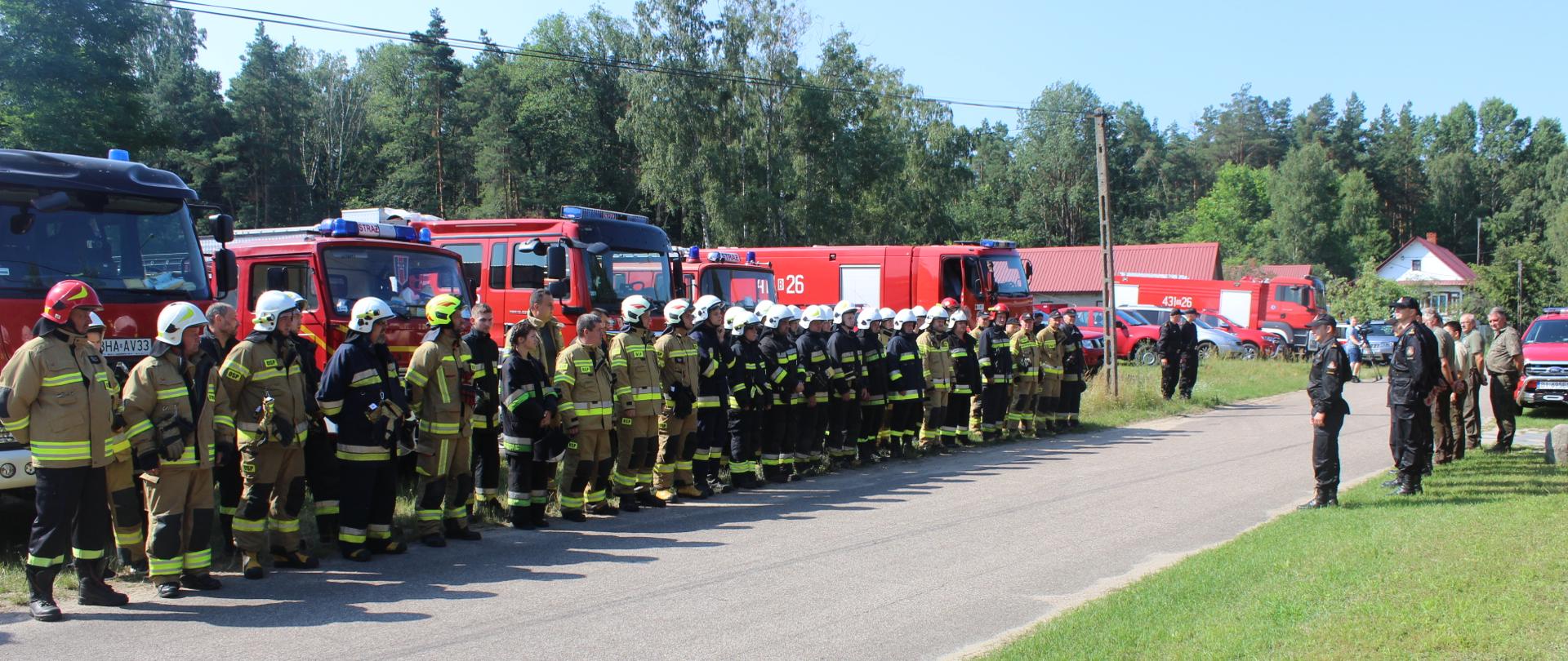 Oficjalne rozpoczęcie ćwiczeń. Strażacy PSP i OSP ustawieni w szyku wzdłuż drogi na tle samochodów ratowniczo-gaśniczych. Naprzeciw nich organizatorzy ćwiczeń - kadra dowódcza KP PSP w Bielsku Podlaskim, KP PSP w Hajnówce oraz przedstawiciele Nadleśnictwa Bielsk