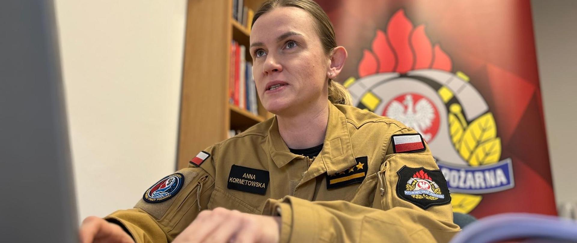 Strażak, kobieta, w mundurze służbowym siedzi za biurkiem, przed komputerem. Ręce trzyma na klawiaturze. Za nią baner reklamowy.