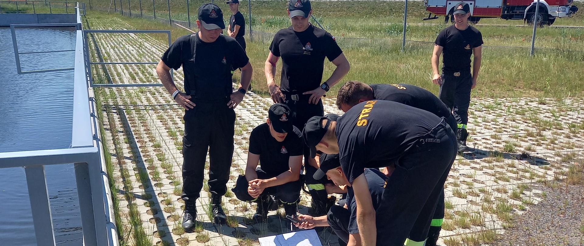 Strażacy w grupie, przy zbiorniku wodnym analizują mapę oraz ustalają położenie i współrzędne wykorzystując GPS. W tle widać drogę S5 oraz pojazd ratowniczo-gaśniczy.