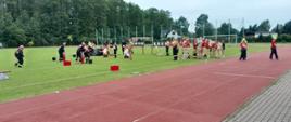 Rozległy widok na boisko sportowe z czerwoną bieżnią na pierwszym planie. Na murawie rozgrywane są konkurencje strażackie. Widać grupy ludzi w różnych strojach (ciemne kombinezony, beżowe i czerwone stroje), w tym osoby przenoszące wiadra z wodą i obsługujące sprzęt gaśniczy. W tle widoczne są drzewa, siatki sportowe i budynki.