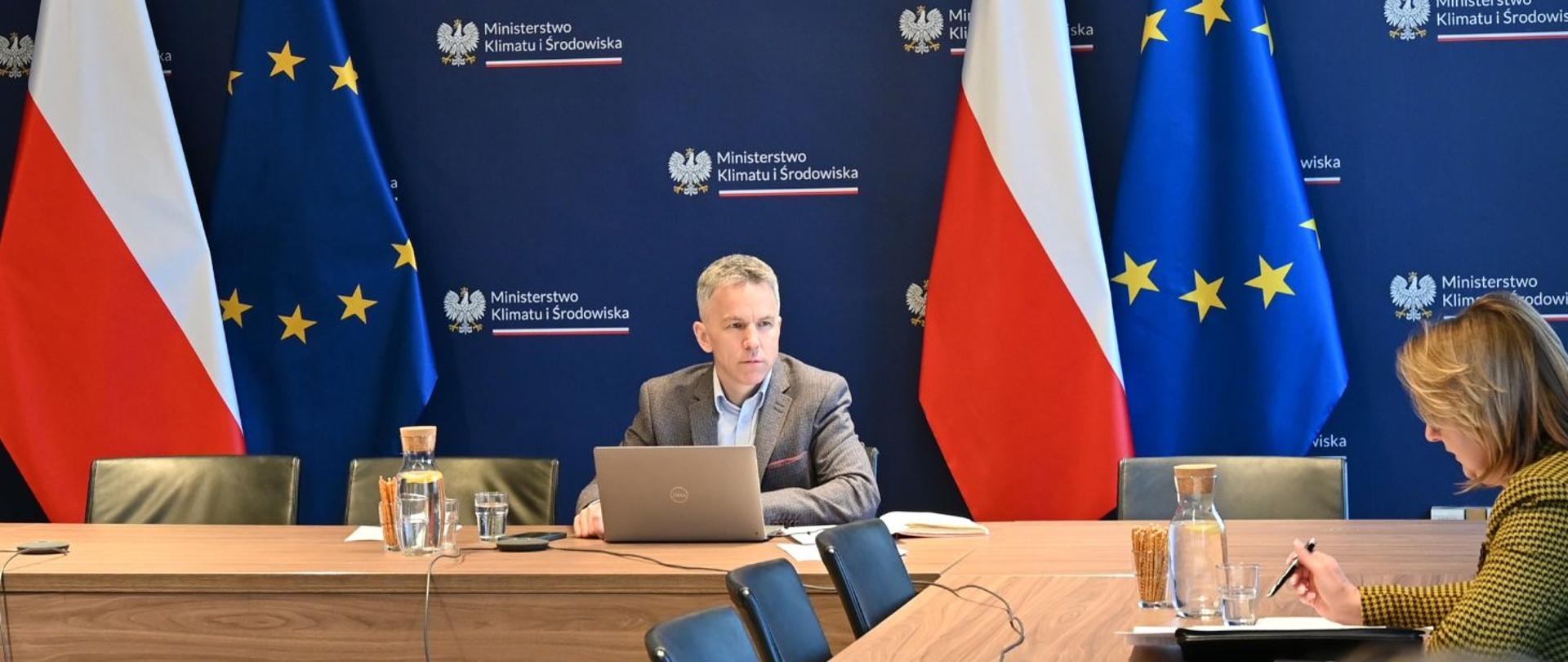 Na zdjęciu widać Szymona Tumielewicza siedzącego przy otwartym laptopie za biurkiem na tle ścianki z logotypem Ministerstwa Klimatu i Środowiska. Po obu stronach ustawione są flagi Polski oraz Unii Europejskiej. Na blacie stoi także karafka z wodą, szklanki oraz paluszki. Po prawej stronie, w rogu zdjęcia, widać kobietę siedzącą bokiem, która spogląda w notatki leżące na stole.