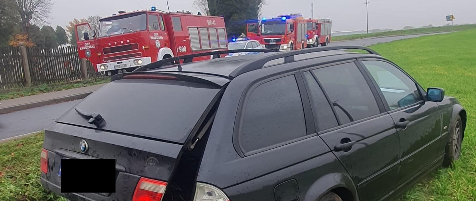 Na łące widać ciemny samochód z uszkodzonym tyłem, w oddali widać trzy samochody straży pożarnej oraz jeden radiowóz Policji